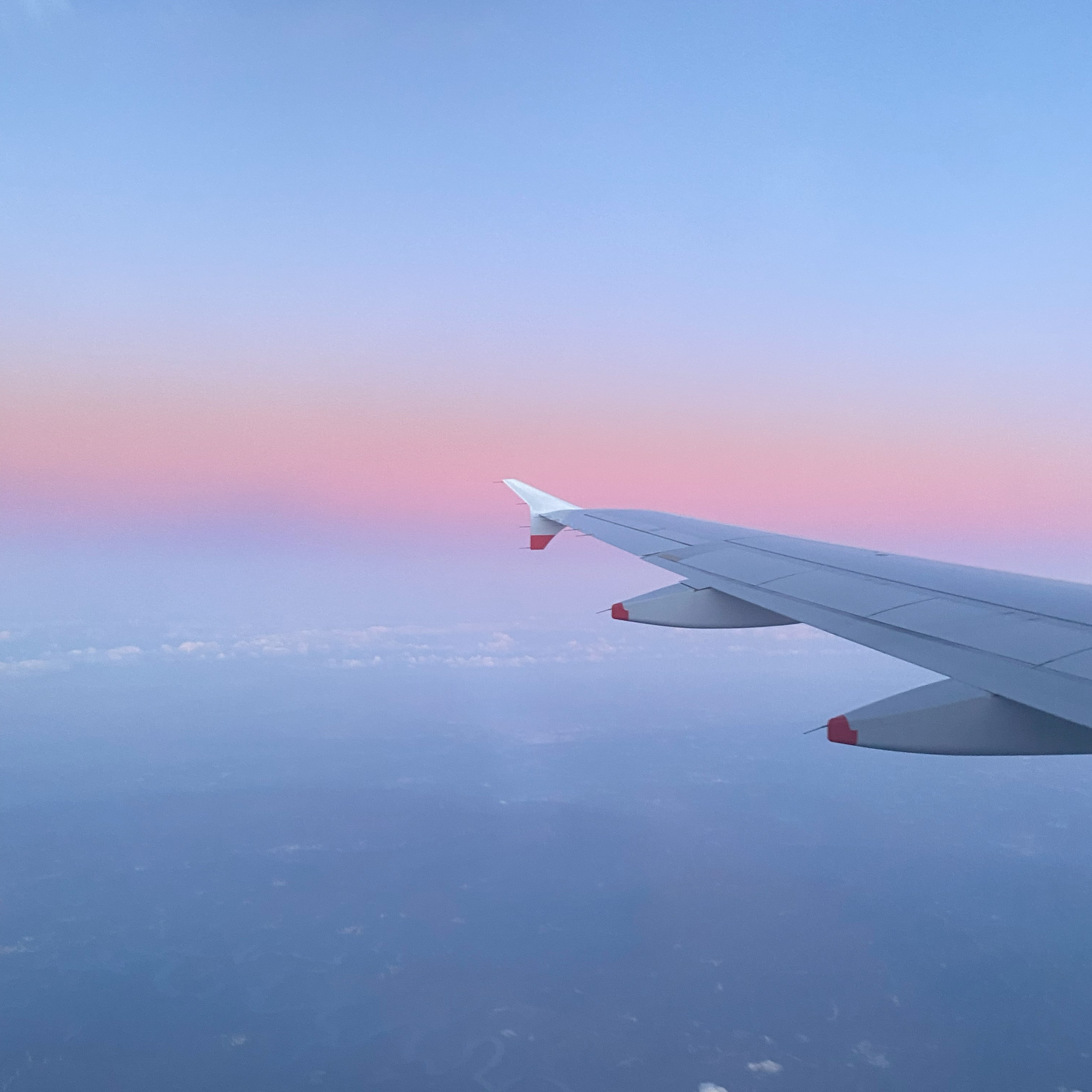 Vista da janela do avião, mostrando a ponta da asa com o céu ao fundo em tons pastel de rosa, roxo e azul durante o nascer ou pôr do sol.
